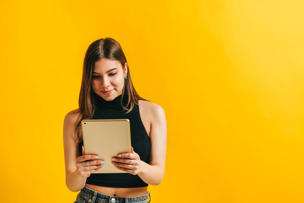 Jolie jeune femme brune utilisant un ordinateur tablette et souriant, isolée sur fond jaune dans un studio avec espace de copie. - Photo, image