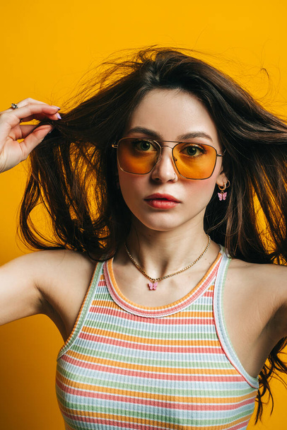 Portrait of attractive stylish woman in sunglasses with long brunette hair isolated on yellow background in studio. - 写真・画像