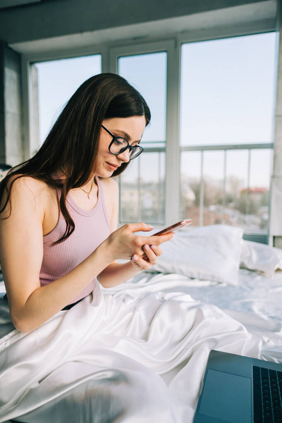 Young caucasian woman resting on a bed at home and using laptop computer and smartphone. Chatting with friends, shopping online.	 - Foto, imagen