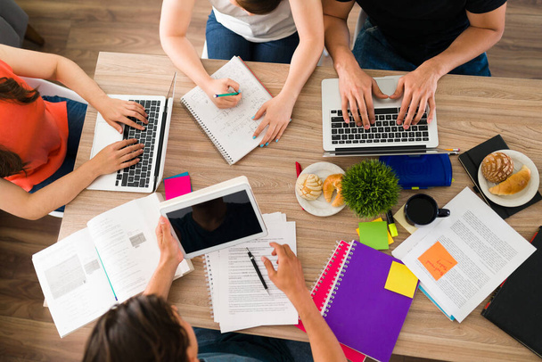 Вид сверху на четырех студентов колледжа, сидящих за столом, выполняя домашнюю работу вместе с книгами, тетрадями и ноутбуками  - Фото, изображение