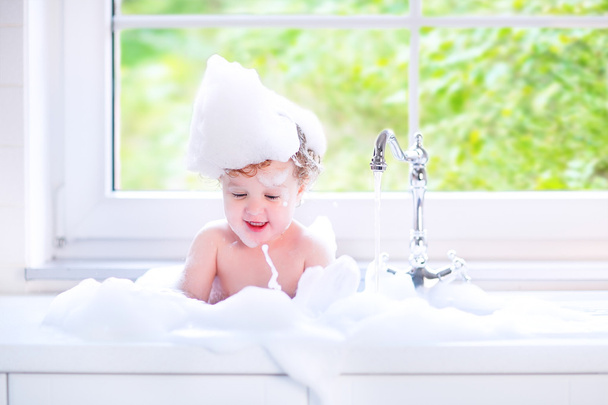 Funny baby girl playing with water and foam in a big kitchen sin - Photo, Image