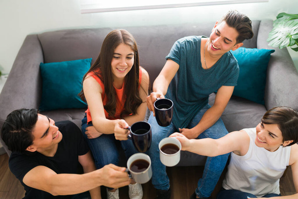 Υψηλή γωνία τεσσάρων ευτυχισμένων φίλων από το κολέγιο που κάνουν πρόποση με φλιτζάνια καφέ και κάνουν μια ωραία συζήτηση στο σαλόνι - Φωτογραφία, εικόνα