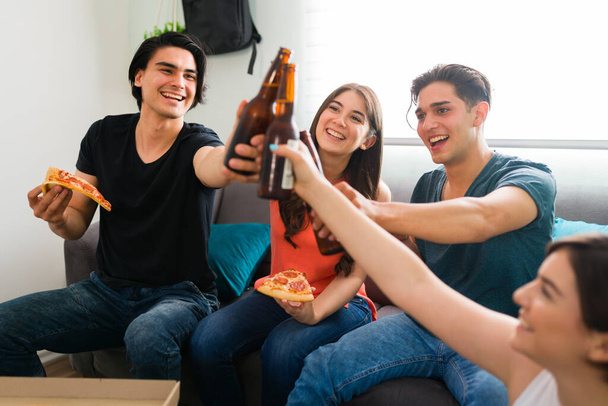 Выпьем за нашу прекрасную дружбу! Молодые друзья из колледжа улыбаются, когда говорят тост за холодным пивом и едят пиццу дома.  - Фото, изображение