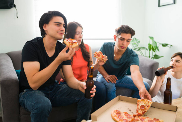 Голодні друзі коледжу насолоджуються піцою та пивом з друзями, сидячи у вітальні вдома
  - Фото, зображення