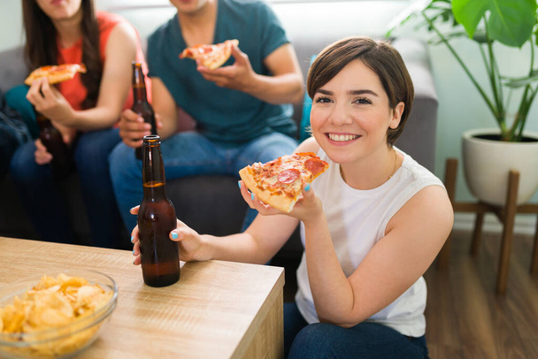 Hermosa mujer joven caucásica sonriendo mientras hace contacto visual y sostiene una rebanada de pizza y una botella de cerveza durante la fiesta de un amigo en casa - Foto, Imagen
