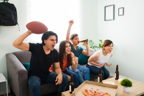 Excited college friends watching a football game and celebrating in the living room because their soccer team is winning - Photo, Image