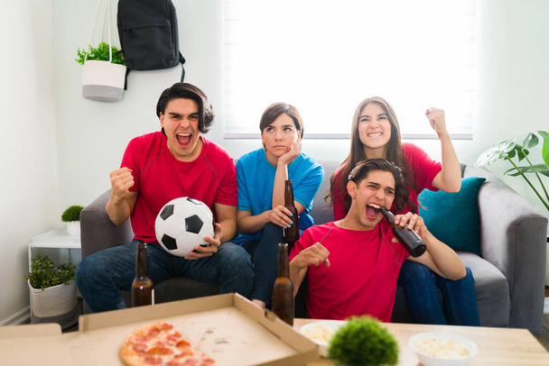 Vrolijke college vrienden dragen voetbal truien en wroeten voor hun favoriete team tijdens het kijken naar de wedstrijd op de woonkamer tv - Foto, afbeelding