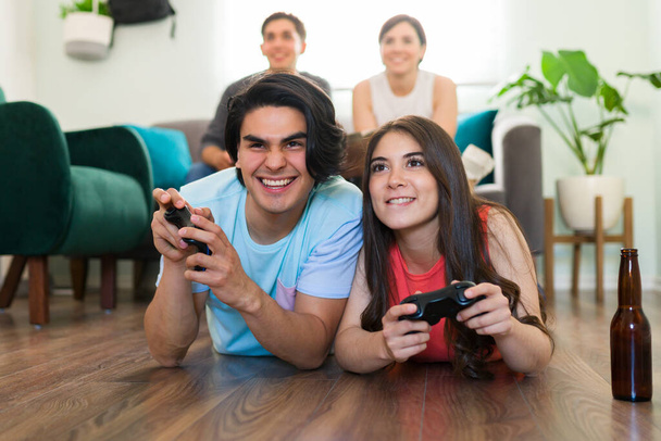 Atractiva pareja tumbada en el piso de la sala de estar mientras se concentran en jugar un videojuego en la televisión con sus amigos - Foto, Imagen