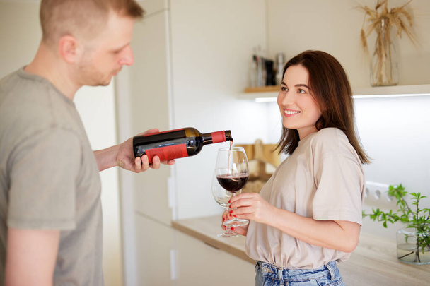 love, dating and romance concept - cute couple in love pouring red wine into glasses in bright kitchen - Foto, Bild