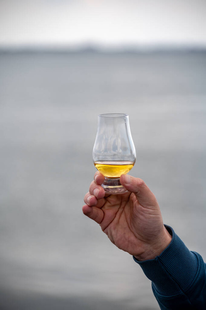 Hand holding dram of single malt scotch whisky on seashore in Scotland - Photo, Image
