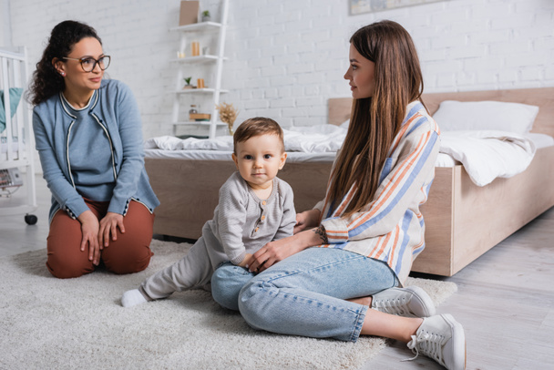 interracial women looking at each other near baby boy sitting on carpet  - Photo, Image