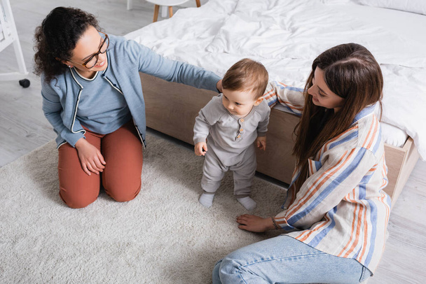 high angle view of interracial women looking at baby boy standing on carpet  - Fotoğraf, Görsel