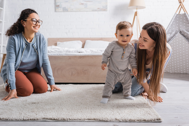 pleased interracial women looking at infant boy standing on carpet  - 写真・画像