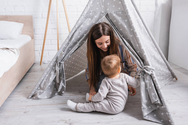 happy mother looking at baby son sitting near tipi in bedroom  - Photo, Image