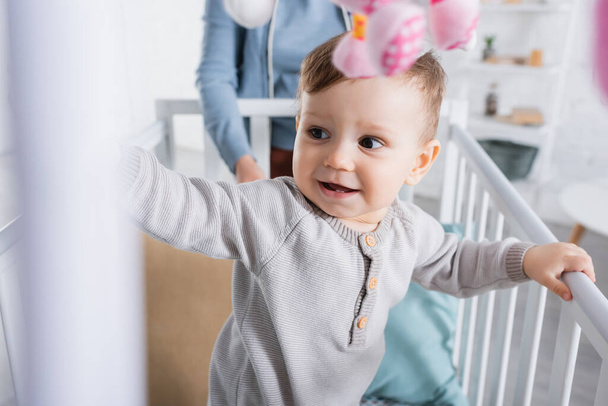 happy infant boy in baby crib with hanging soft toy on blurred foreground - Zdjęcie, obraz