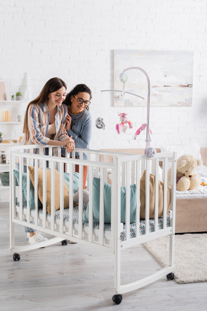 happy interracial women looking at infant boy in baby crib in bedroom  - Fotó, kép
