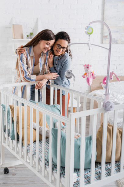 smiling interracial women looking at infant boy in baby crib in bedroom  - Photo, Image