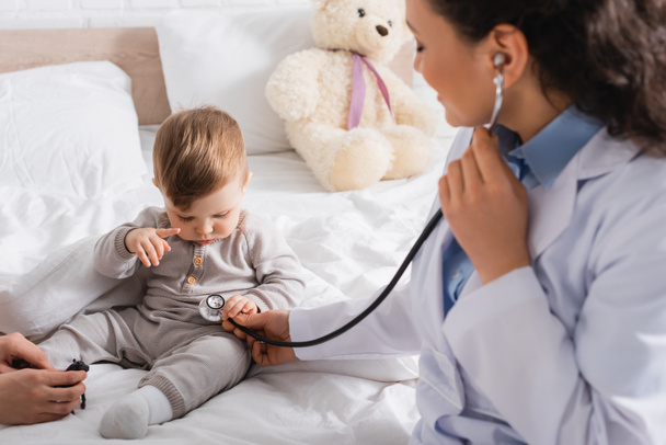 blurred african american pediatrician in white coat examining baby boy with stethoscope near mother - Φωτογραφία, εικόνα