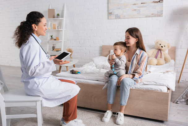 happy african american pediatrician in white coat holding digital tablet with blank screen near mother and son in bedroom  - Photo, image