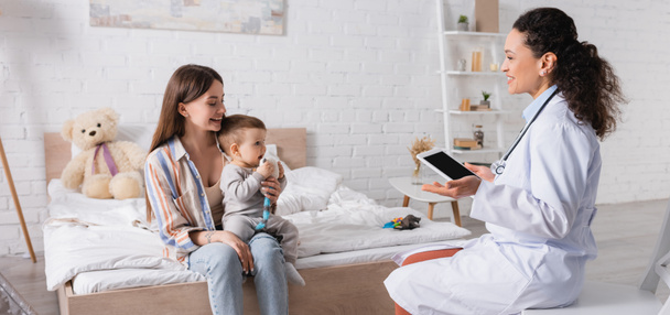 pediatra afroamericano de capa blanca sosteniendo tableta digital con pantalla en blanco cerca de madre feliz e hijo en el dormitorio, pancarta - Foto, imagen
