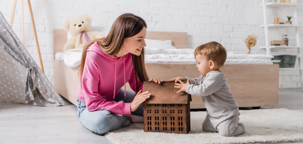 Mutlu anne, oyuncak evin yanındaki halının üstünde oturan oğluna bakıyor. - Fotoğraf, Görsel