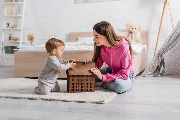 happy mother looking at infant son sitting on carpet near toy house - Foto, Imagen