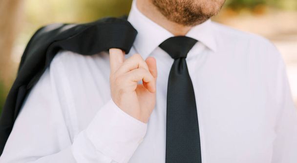Beyaz gömlekli ve siyah kravatlı bir adam dikiliyor ve omzunun üzerinden ceketini tutuyor, yakın plan.  - Fotoğraf, Görsel