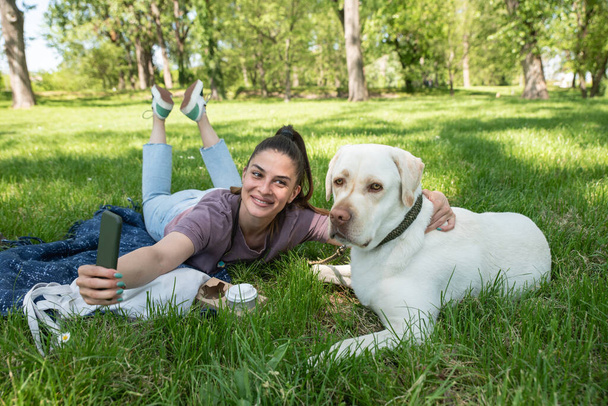 Young beautiful woman lying in the park with her pet dog a golden Labrador Retriever and takes a selfie photo with her smartphone while having fun and enjoying a beautiful sunny day - Foto, Imagen
