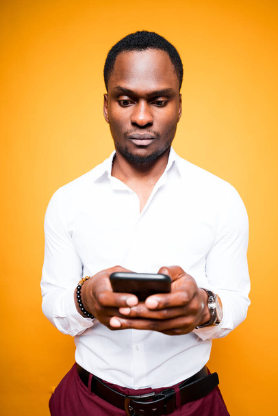 Handsome dark-skinned guy in a white shirt works on the Internet while holding a phone in his hands - Фото, зображення