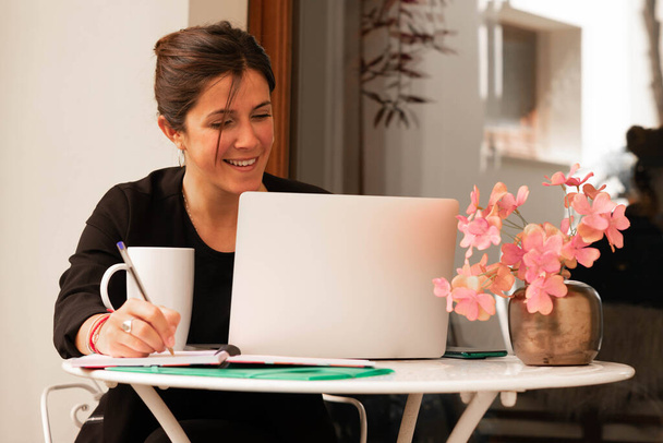 vízszintes kép egy fiatal nővel, aki otthon dolgozik, laptoppal mosolyogva egy megbeszélésen, miközben jegyzetel - Fotó, kép