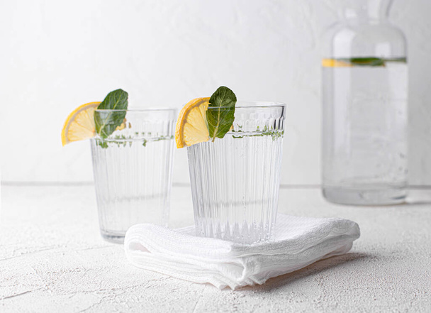 Due bicchieri di acqua dolce, decorati con spicchi di limone e foglie di menta su un tavolo bianco. Concentrazione selettiva. Vista frontale - Foto, immagini