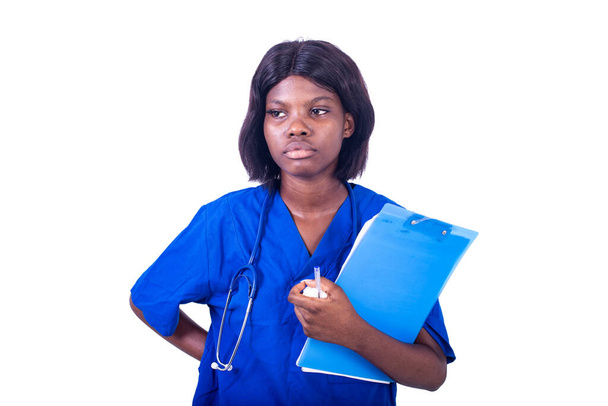 νεαρή νοσοκόμα με στολή στέκεται σε λευκό φόντο με στηθοσκόπιο κοιτάζοντας μακριά, ενώ μεταφέρουν ένα σημειωματάριο. - Φωτογραφία, εικόνα