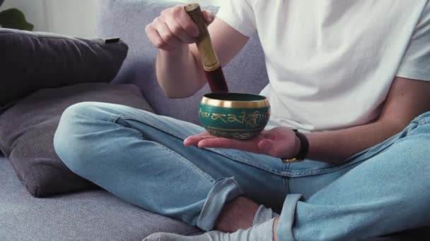 Боротьба з депресією і стресом. Молодий чоловік використовує тибетську чашу для співу у вітальні. Letters mean tibetan mantra: Om Mani Padme Hum, Слава коштовності в The Lotus - Кадри, відео