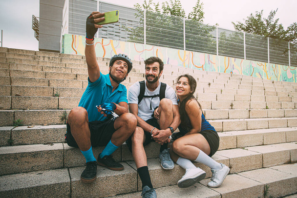 Grupa szczęśliwych, uśmiechniętych przyjaciół ubranych w strój sportowy, siedząca na miejskich schodach i robiąca selfie telefonem komórkowym. Przyjaciele robiący zdjęcie ze smartfonem w miejskim otoczeniu. tło z copyspace. Pojęcie wolnego czasu. Koncepcja przyjaźni. - Zdjęcie, obraz