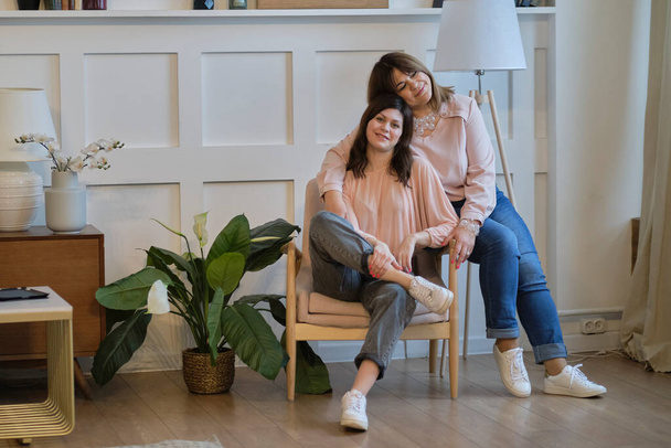 Beautiful senior mom and her adult daughter are hugging and smiling while sitting on couch at home - Zdjęcie, obraz