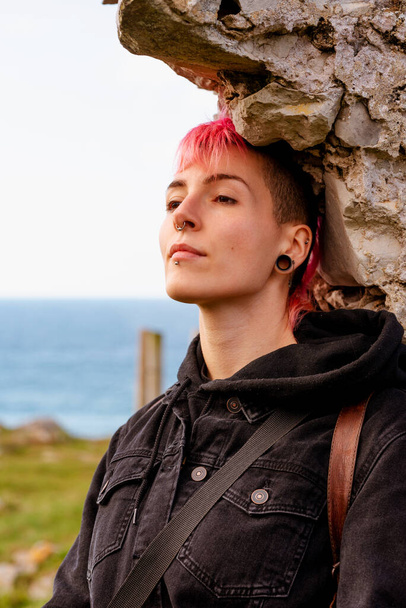 portrait of an androgynous woman leaning against a stone wall, gazing into infinity with a serene gesture and without makeup. gender and identity. young girl with pink hair, mohawk, and piercings. - Photo, image