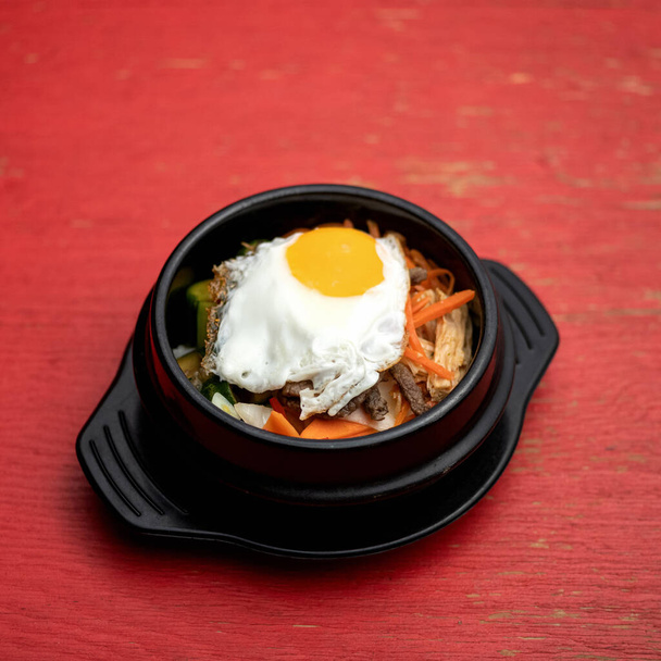 Pan-Asian cuisine. Black clay pot with food on red background. Dish of vegetables and meat, garnished with fried egg on top. Spicy oriental dish - Photo, Image