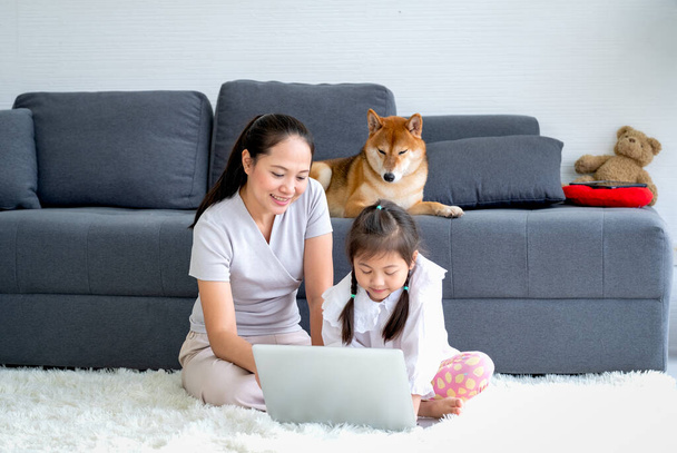 Asiatische Mutter und kleines Mädchen schauen auf Laptop und ihr Shiba-Hund bleibt auf dem Sofa und interessiert sich auch für Laptop, indem er auf den Monitor schaut. Konzept einer glücklichen asiatischen Familie, die während einer Pandemie zusammenbleibt. - Foto, Bild