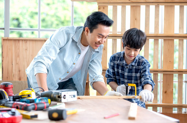 Asiatische Vater schauen sorgfältig Sohn verwenden Säge mit Holz in ihrem Arbeitsplatz der Tischlerei mit glücklichen Gefühl. Asiatisches Familienkonzept, zu Hause zu bleiben und gemeinsam gutes Beziehungshobby zu genießen. - Foto, Bild