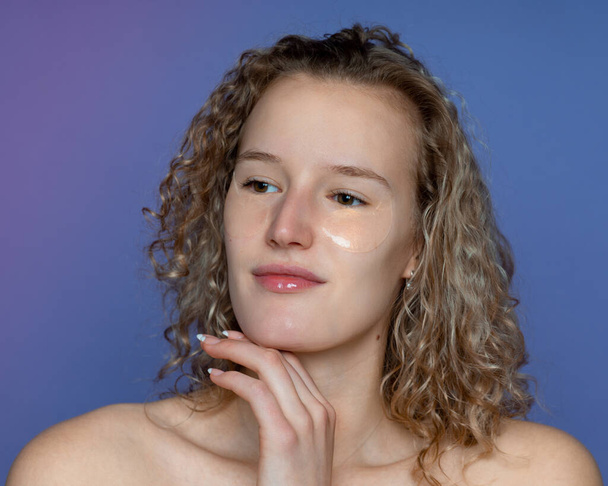 inspired young beautiful girl with cosmetic patches for the area around the eyes, cleansing, skin improvement, looking to the lenses, with curly hair, acne is visible on the skin, blue background - Fotó, kép