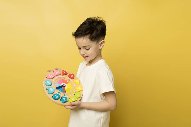 Little boy studies with wooden clock, portrait on yellow studio background. Child learns to tell time, carefully looks at the dial. Concept of learning, games for the development of preschool children - Photo, Image