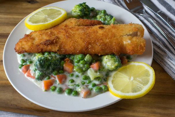 Paniertes Fischfilet mit gemischtem Gemüse und Bechamelsauce heiß auf einem Teller mit Zitronenscheiben serviert - Foto, Bild
