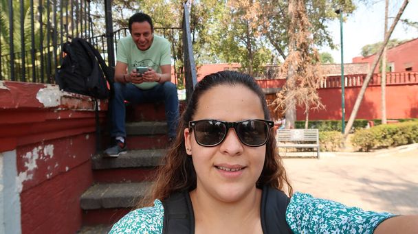 junge Frau mit dunkler Brille macht Selfie und im Hintergrund ist ein Mann zu sehen - Foto, Bild