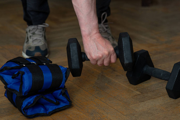 Equipment for exercising the muscles of the arms and legs. Weights and sandbags on the wooden floor. The man takes a dumbbell and picks it up with one hand - Photo, Image