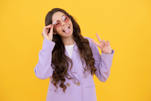 beauty and fashion. female fashion model. pretty look of young girl in jacket. portrait of happy child on yellow background. express positive emotions. cheerful kid with curly hair in sunglasses. - Photo, Image