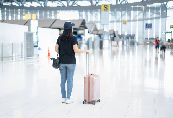 Η πίσω όψη της Ασιάτισσας που μεταφέρει βαλίτσα αποσκευών και φοράει χειρουργική μάσκα ετοιμάζεται να κάνει check-in για να πάρει την κάρτα επιβίβασης στο αεροδρόμιο. Κατά τη διάρκεια της επιδημίας Covid-19.  - Φωτογραφία, εικόνα