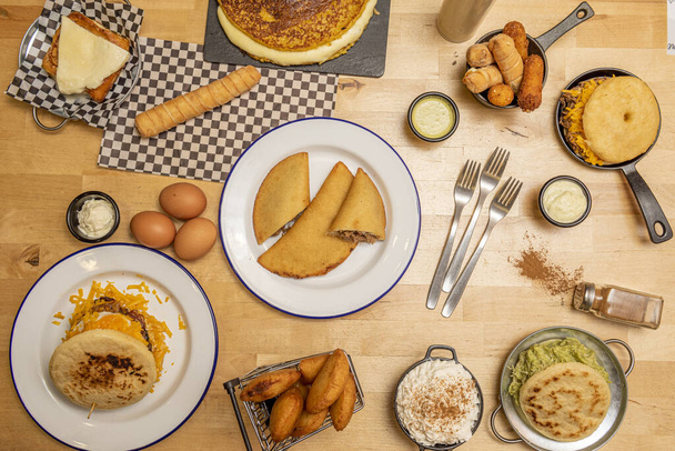 Типичные тарелки венесуэльской еды на светлом деревянном столе. Арепас, качапа, текео, кукурузные эмпанады, яйца и соусы - Фото, изображение