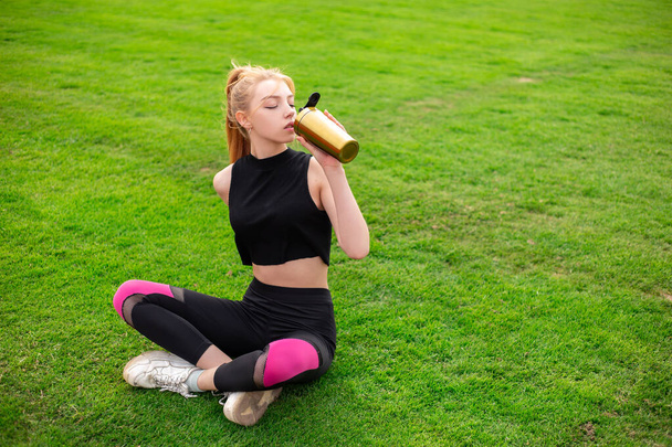 Ένα κορίτσι κάθεται σε στάση λωτού στο γήπεδο ποδοσφαίρου, κοιτάζει την κάμερα και χαμογελάει. Αθλητική γυναίκα σε αθλητικά ρούχα στο πράσινο γκαζόν ή γρασίδι με χώρο αντίγραφο. Μεταλλικό μπουκάλι αναδευτήρα - Φωτογραφία, εικόνα
