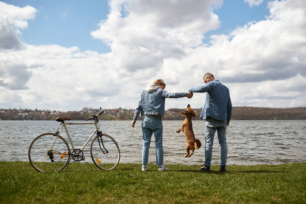 Young couple on a bicycle resting by the lake playing with their pet. - Photo, Image