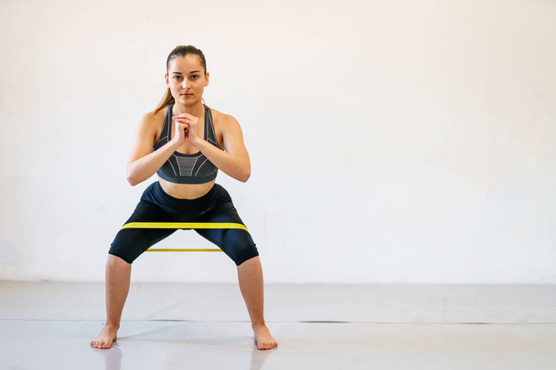 Giovane donna caucasica istruttrice di fitness formazione di fronte alla parete bianca con elastico di resistenza - full length femminile sportiva con spazio copia - Foto, immagini
